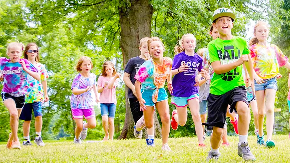 Smiling kids in colorful camp shirts running through a grassy field, used as both the featured header for SpringHill Mini and the card image on the /camps program overview page